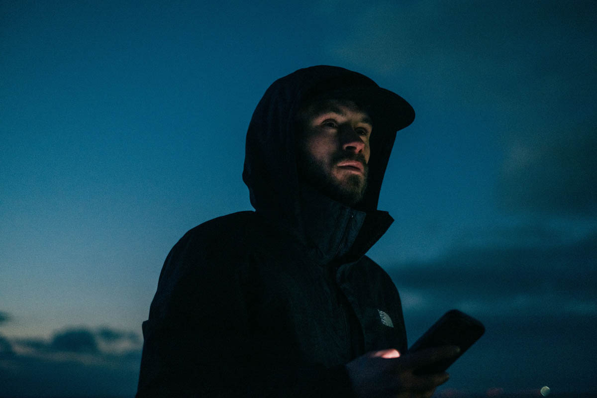Person i mörk jacka med huva står utomhus i skymning och tittar på en mobiltelefon, med mörkblå himmel och moln i bakgrunden.