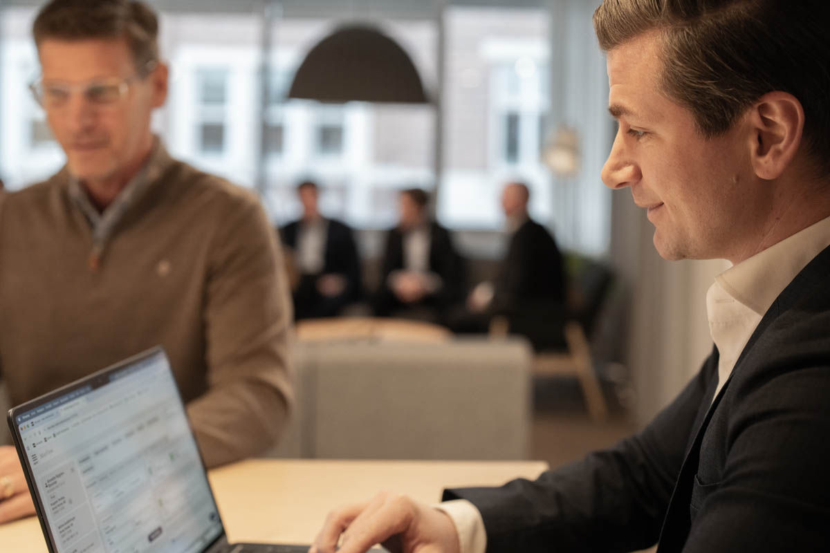 Person arbetar vid laptop i kontorsmiljö medan en kollega står bredvid och diskuterar, med fler personer i bakgrunden