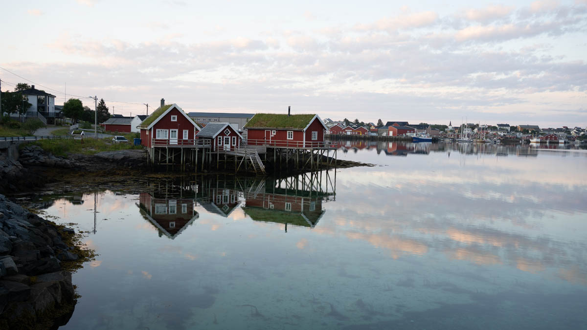 Röda sjöbodar på pålar vid stilla kust med spegling i vattnet, skärgårdsbebyggelse i bakgrunden