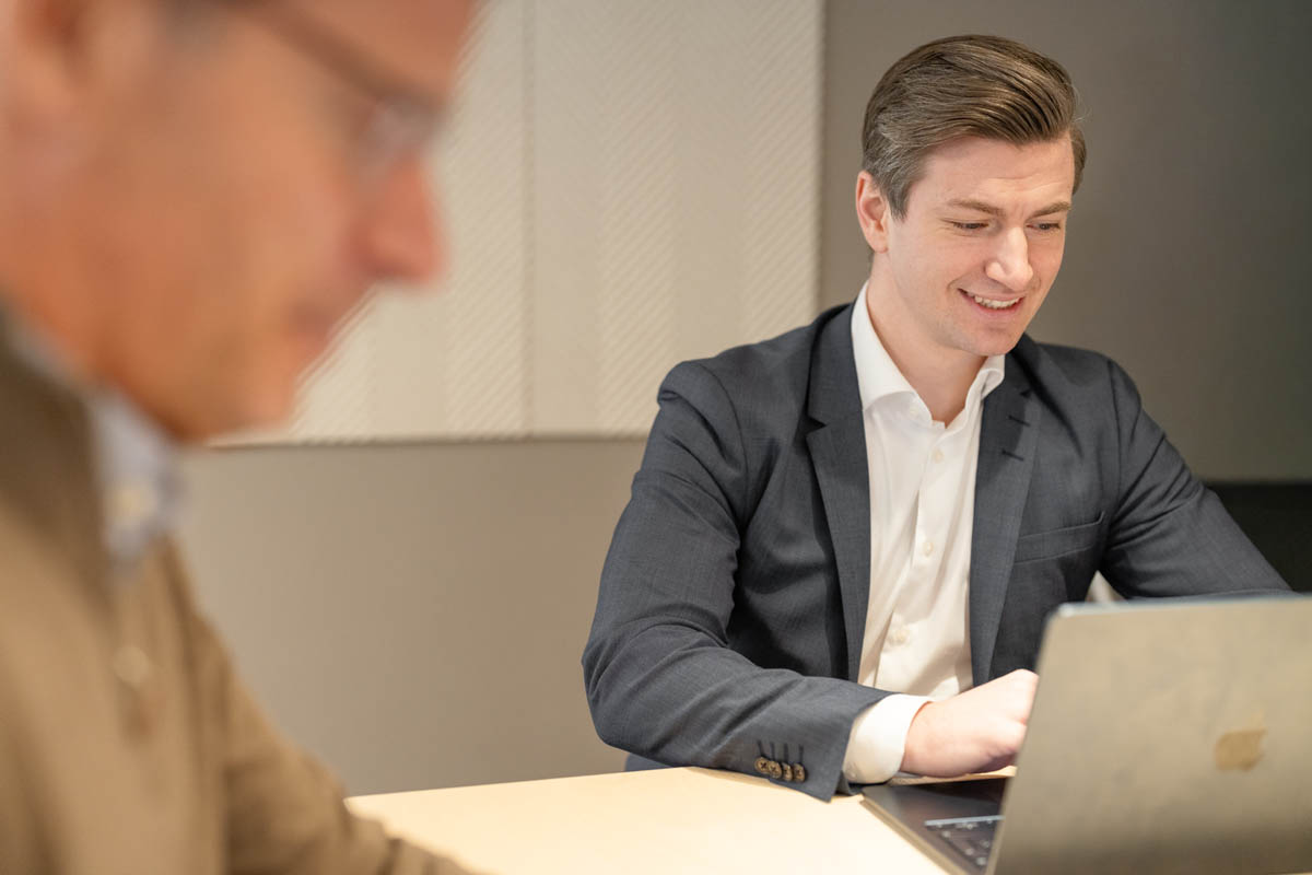 Person sitter vid bord och arbetar på laptop i mötesrum, med en annan person i förgrunden